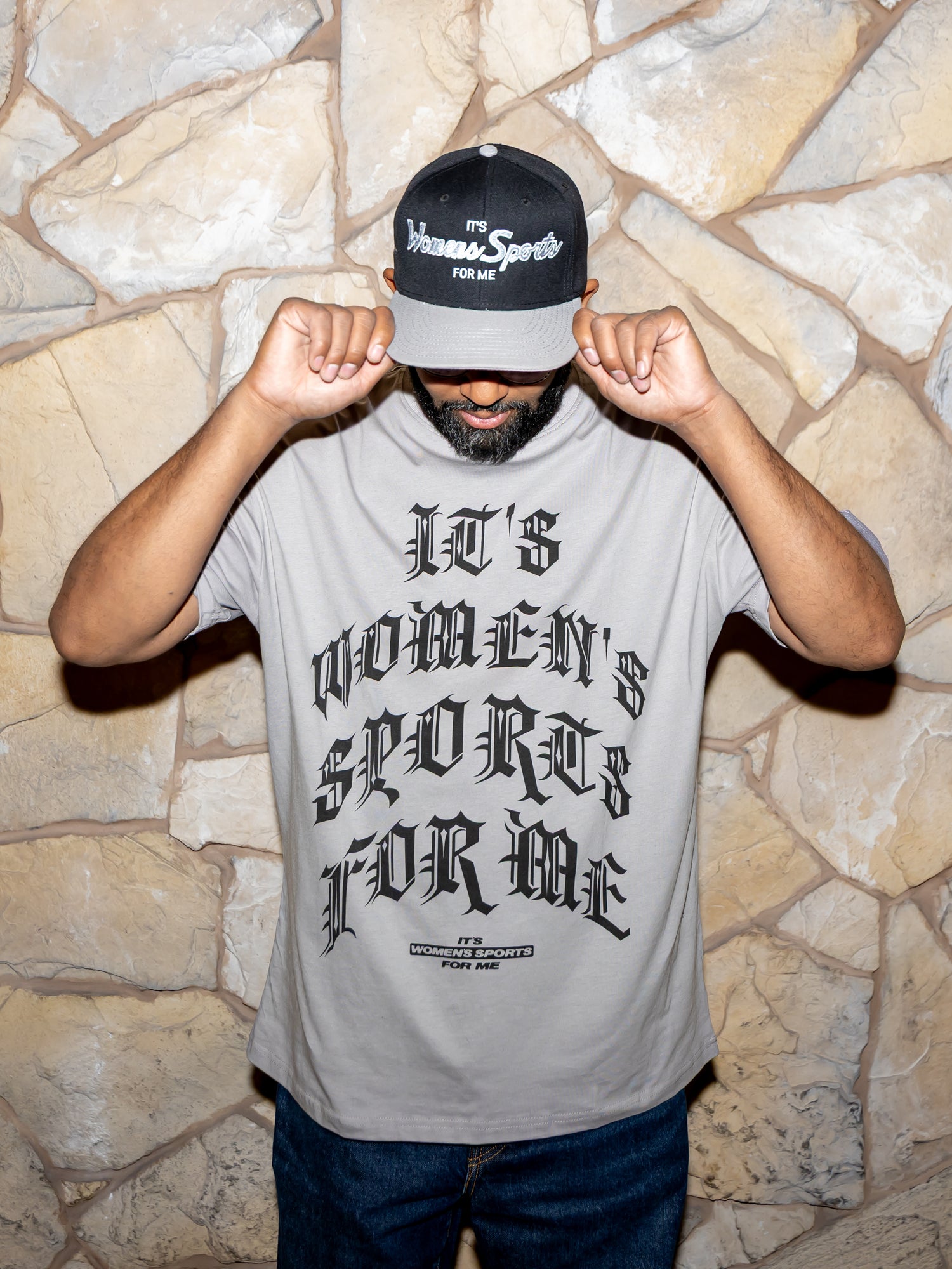 Person wearing a gray t-shirt with text and a cap, standing against a stone wall.